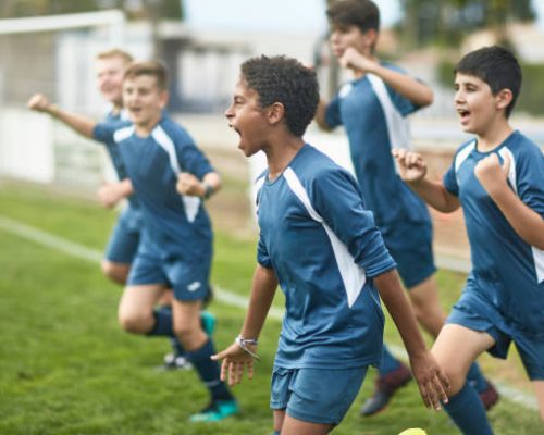 Energetic preteen and teenage male footballers cheering and punching the air as they run onto field for training session.